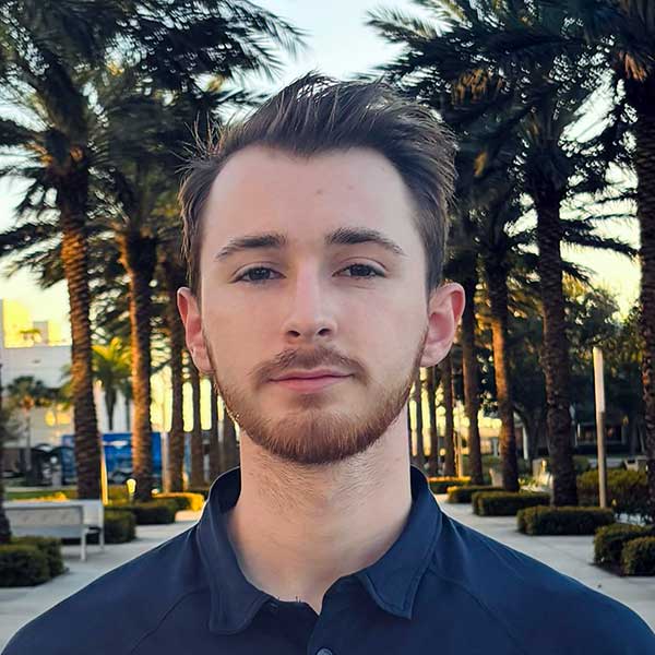 Conor, with light skin tone, looks calmly at the camera, standing centered in front of palm-tree lined Legacy Walk.