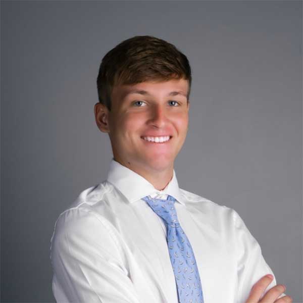 Jonathan wears a white button down shirt and light blue tie, smiling, arms folded.