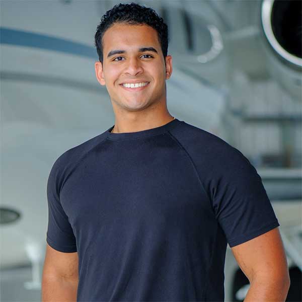 Renso wears a dark blue t-shirt and smiles confidently into the camera, standing in front of an airplane. He has medium skin tone.