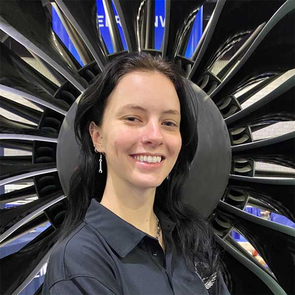 Shelby, with light skin tone, in a grey button down shirt, stands in front of an engine turbine fan.