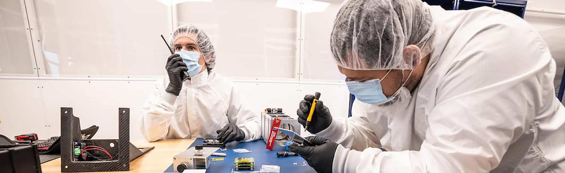 Two students wearing clean room suits work on satellite in lab.