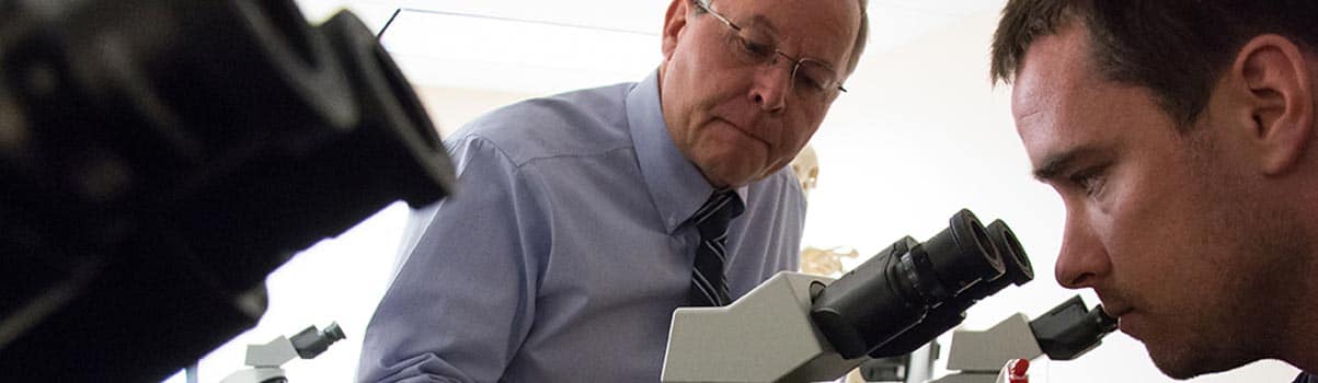 a student looks into a microscope while a professor watches