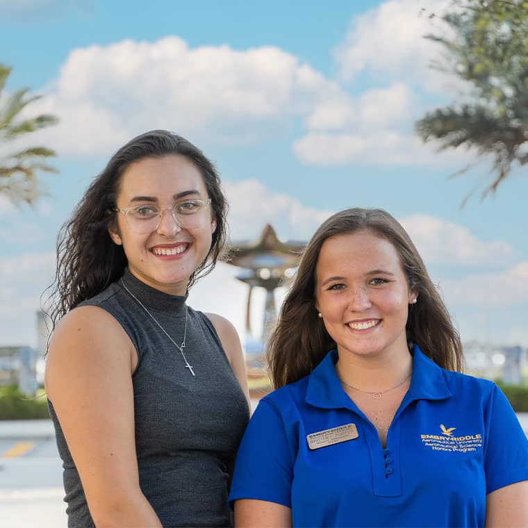 Two students, one with an Embry-Riddle name tag, pose smiling in front of the Pathways to the Sky sculpture on the Daytona Beach Campus.