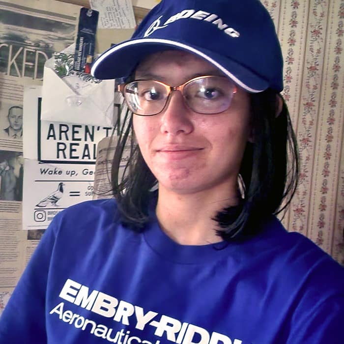 A student with light skin tone wearing glasses, Boeing cap, and Embry-Riddle t-shirt. Behind her is a flyer with the slogan for the satirical conspiracy group, Birds Aren't Real.