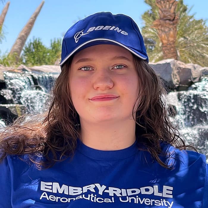 Student with medium length brown hair looks into the camera - she's wearing a blue Boeing baseball cap and blue Embry-Riddle t-shirt and standing in front of a waterfall.