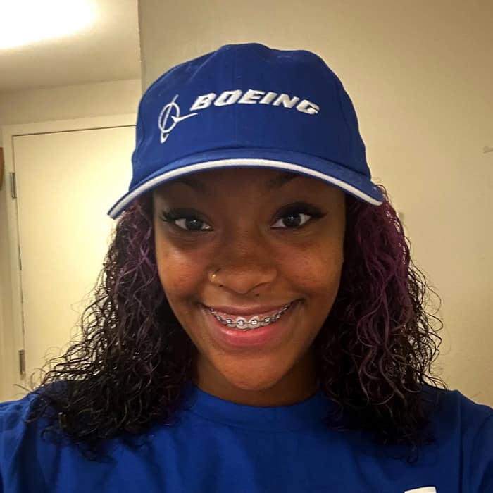 Black woman with medium length brown hair smiles into the camera, wearing a blue Boeing cap.
