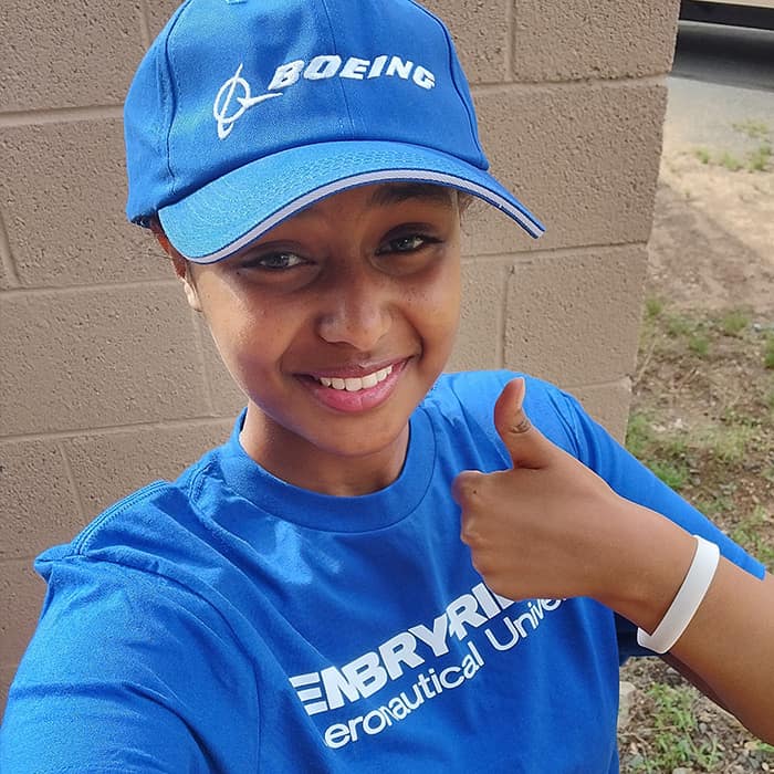 Liyat has dark skin tone; she gives a thumbs up, smiling into the camera. She's taking a selfie outside and wearing a Boeing cap and Embry-Riddle t-shirt.