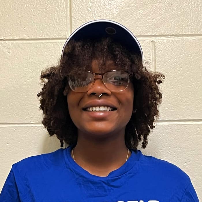 A black woman with natural curls wearing glasses and a cap smiles into the camera.