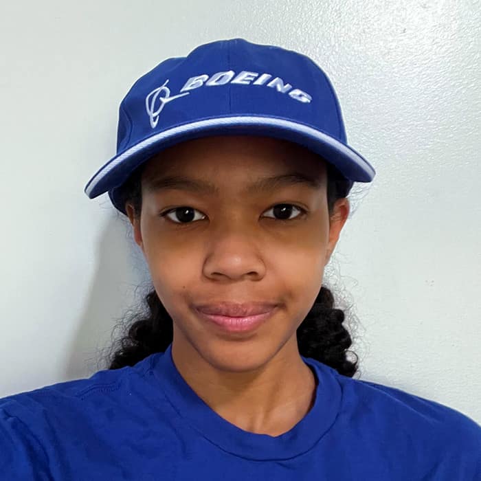 A young Black woman with her hair pulled back wears a blue Boeing cap.