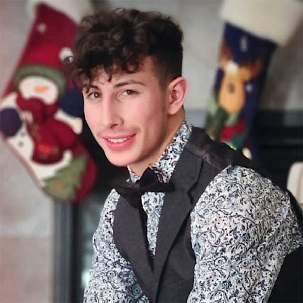 Dominic, with curly hair, a patterned shirt and vest, and a bow tie, poses in front of a fireplace mantel decorated with holiday stockings.