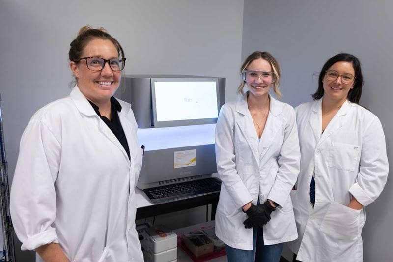 Three female scientists in lab coats stand in front of imaging machine