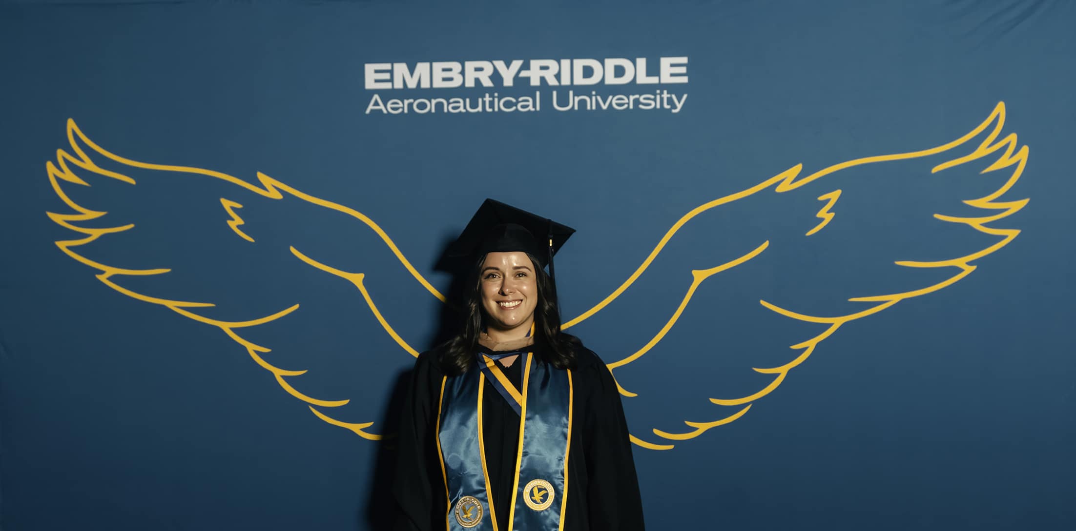 Woman in cap and gown with eagle wings on backdrop behind her. 